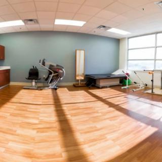 A spacious physical therapy room equipped with exercise machines and treatment tables, brightly lit by natural daylight.