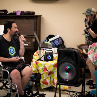 A lively indoor performance featuring a man in a wheelchair singing while another person looks on, surrounded by musical equipment.