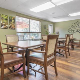 A bright dining room with wooden tables and chairs, featuring artwork on the walls and natural light from a large window.