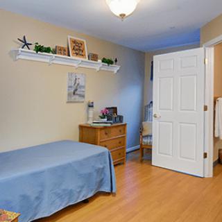 A cozy bedroom featuring a single bed with blue bedding, a wooden dresser, and a shelf above.
