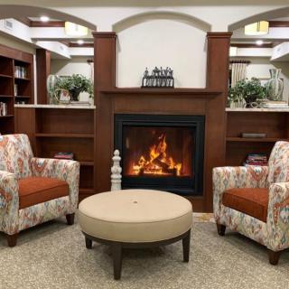 A cozy living room with a fireplace, two patterned chairs, and a round table, surrounded by bookshelves.