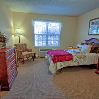 A cozy bedroom with a bed, dresser, and chair, softly lit by a lamp near the window.