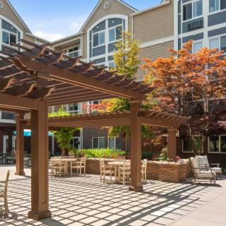 A sunny outdoor patio area featuring a wooden pergola, tables, and chairs, with an apartment building and trees in the background.