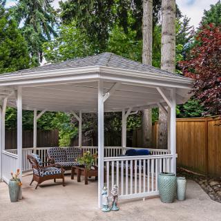 A cozy outdoor gazebo with comfortable seating, surrounded by lush trees and a privacy fence.