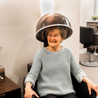 An elderly woman smiles while sitting under a hair dryer at a salon.