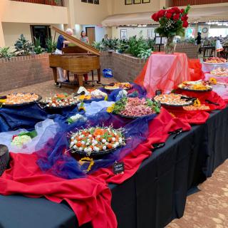 An elegant buffet setup featuring a variety of dishes, with vibrant red and blue drapery, accompanied by a grand piano in an indoor setting.