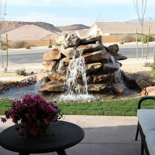 A peaceful garden setting featuring a cascading rock waterfall with a view of distant hills and a cozy patio area.