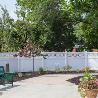A serene outdoor space featuring a green bench, neatly landscaped plants, and a white fence surrounded by trees.