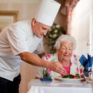 A chef in a white uniform serves a meal to an elderly woman sitting at a restaurant table.