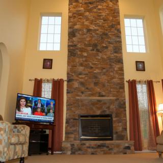 A cozy living room featuring a tall stone fireplace and large windows with light streaming in.