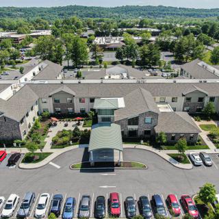 An aerial view of a complex with multiple buildings and a parking lot full of cars, surrounded by greenery.