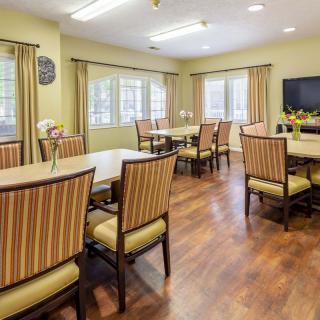 A well-lit dining room with multiple tables and chairs, featuring large windows and wooden flooring.