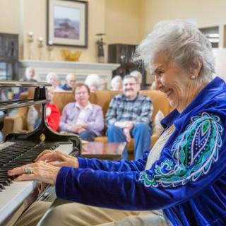 An elderly woman joyfully playing the piano for an attentive audience.
