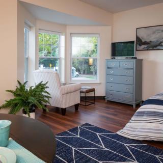 A cozy bedroom with a large window, featuring a comfortable chair, dresser, and decorative plant.