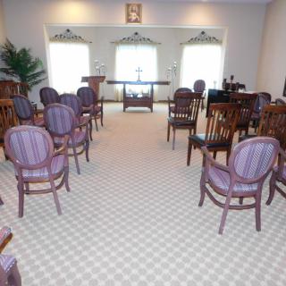 An interior view of a small chapel with wooden chairs, a carpeted floor, and a table at the front.