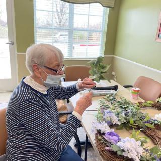 An elderly person wearing a mask is engaged in crafting with flowers at a table indoors.