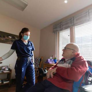 A nurse wearing a medical mask converses with an elderly patient in a home care setting.