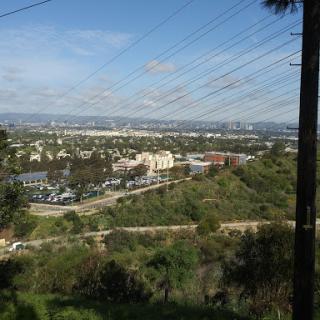 A panoramic view of a cityscape framed by hills and greenery, with power lines crossing the scene under a partly cloudy sky.