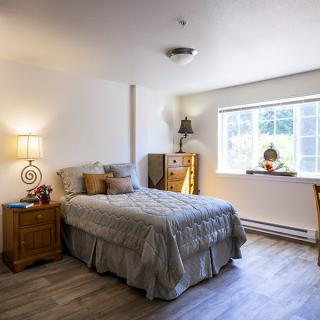 A cozy bedroom with a bed, side table, and chest of drawers illuminated by natural light from the window.