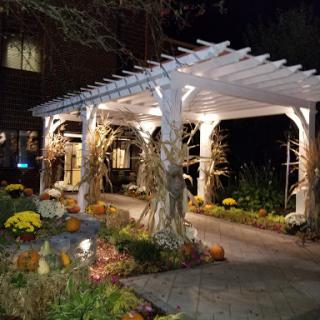 A beautifully lit garden entrance featuring a white pergola adorned with corn stalks and vibrant fall flowers.