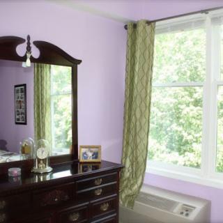 A cozy bedroom featuring a dark wooden dresser, a large mirror, and flowing green curtains by the window.