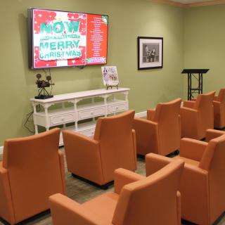 A cozy waiting room with orange chairs, a large screen for presentations, and a green accent wall.