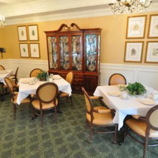 An elegant dining room featuring round tables set with white tablecloths, surrounded by classic chairs and a wooden cabinet against the wall.