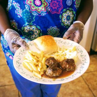 A person holding a plate with meatballs, fries, mashed potatoes, and a bread roll.
