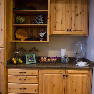 A kitchen area featuring wooden cabinets with open shelves, a countertop with fruit and a drinks dispenser.