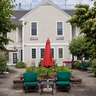 A cozy patio area with green chairs and a red umbrella, set in front of a charming house surrounded by a garden.