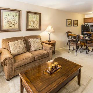 A cozy living room featuring a sofa, wooden coffee table, and artwork on the walls, with a dining area in the background.
