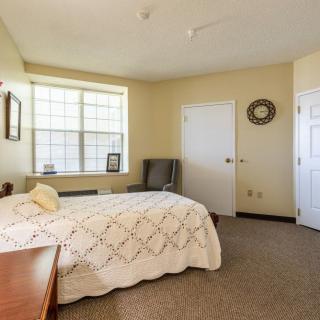 A cozy bedroom interior with a neatly made bed, armchair, and a window allowing natural light.