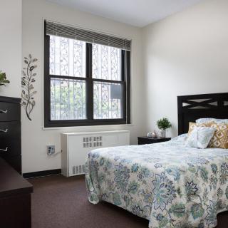 A neatly arranged bedroom featuring a bed with a floral patterned bedspread and a large window allowing natural light to fill the room.