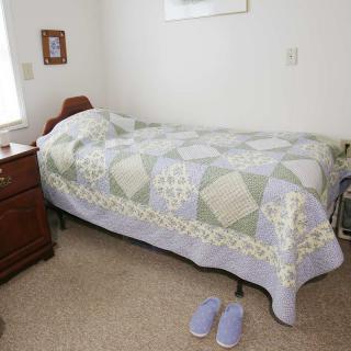 A cozy bedroom with a single bed, quilt, nightstand, and slippers on the carpet.