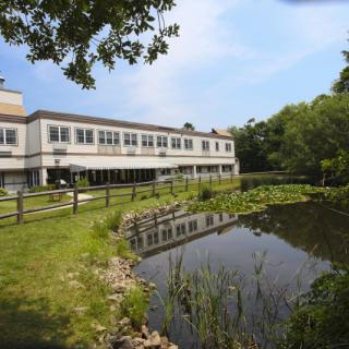 A scenic view of a building by a tranquil lake with lush greenery and clear blue sky.