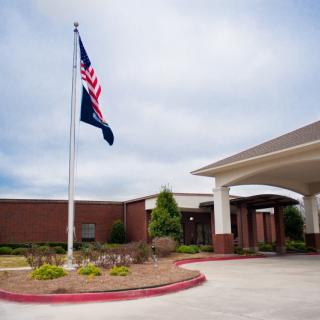A view of a building entrance with a flagpole, featuring a well-maintained landscape and a clear sky in the background.