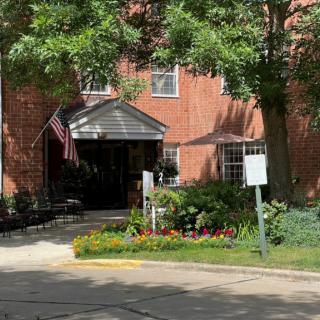 A brick building entrance surrounded by a small garden and trees, with an American flag displayed.