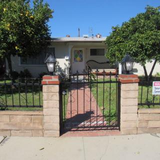 A single-story residential house with a fenced yard and gate, surrounded by trees.