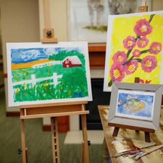 A collection of colorful paintings displayed on easels indoors, featuring a landscape scene and floral artwork.