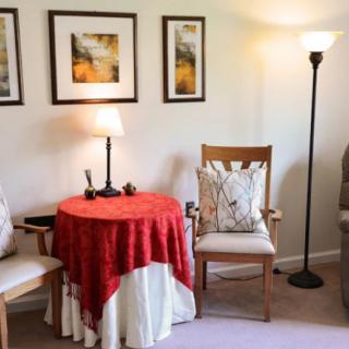A cozy living room corner featuring a table draped with a red cloth, surrounded by wooden chairs, a floor lamp, and framed wall art.