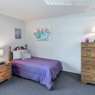 A cozy bedroom with purple bedding, a wooden dresser, and a nightstand adorned with decor.