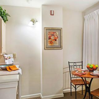 A cozy kitchen and dining area with a small table set for two, featuring a fruit bowl and a framed artwork on the wall.
