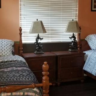 A cozy bedroom features twin beds with decorative lamps on a shared nightstand beneath a window with blinds.