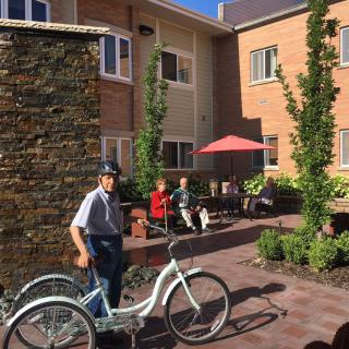 A man with a tricycle in a sunny courtyard area, with people sitting nearby.