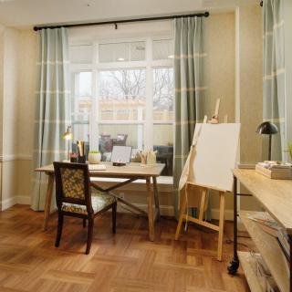 A cozy home office with an art easel by the window, featuring a desk, chair, and soft curtains.