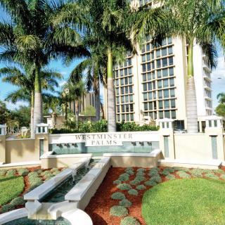 A modern building surrounded by palm trees and neatly landscaped gardens with a fountain in front.