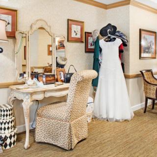 A cozy dressing room featuring a wedding dress on display next to an elegant vanity table with a wicker chair.