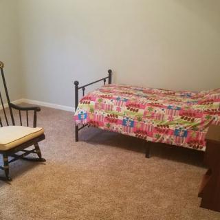 A minimalist bedroom with a single metal-framed bed covered in colorful bedding beside a wooden rocking chair on a carpeted floor.