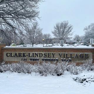 A snowy scene featuring the entrance sign to Clark-Lindsey Village surrounded by trees.