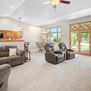 A spacious living room with modern decor, featuring sofas and recliners, a ceiling fan, and large windows allowing natural light.